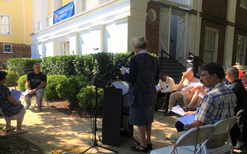 Photo: A woman stands behind a podium reading to a group sitting in chairs. 